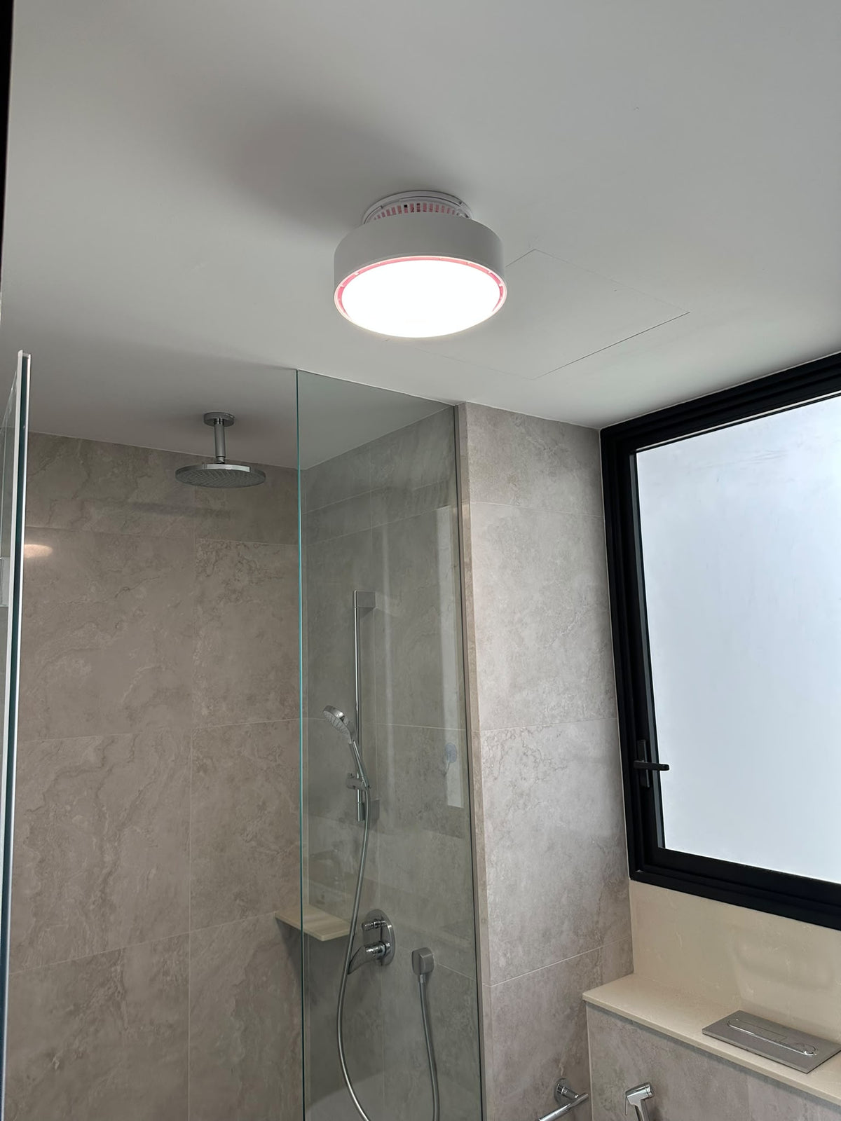 ALANTZ Aura-Flow Compact ceiling fan with its LED light on in a pale stone tiled bathroom beside a glass shower screen